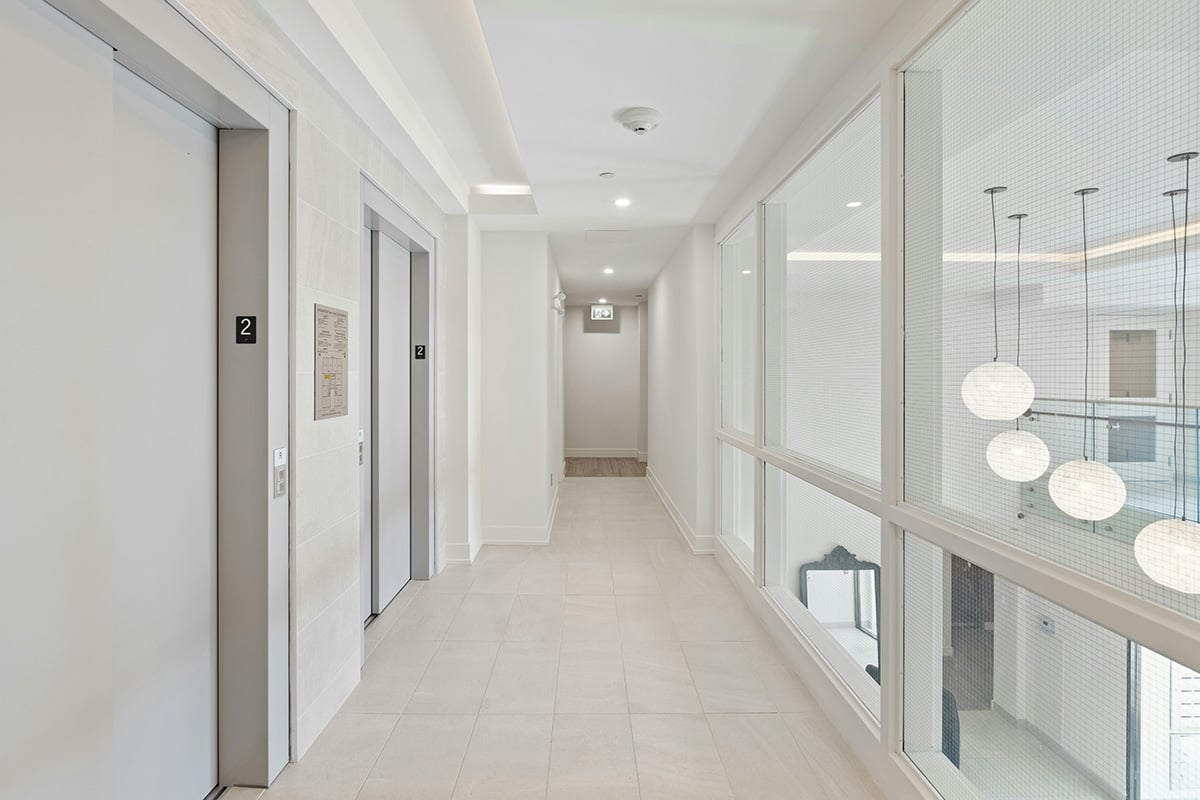 The elevator bank and hallway of an apartment building with glass windows overlooking the lobby
