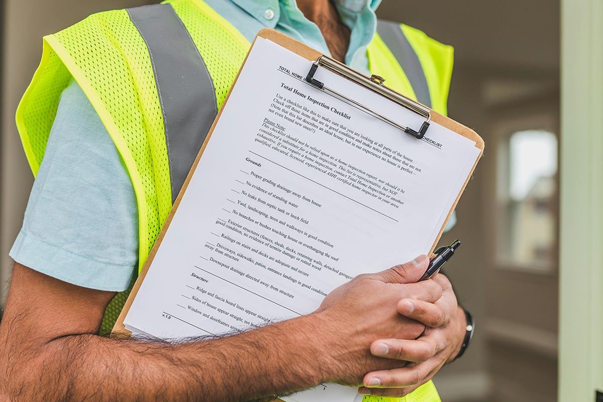 A home inspector in a yellow vest carrying an inspection checklist on a clipboard.