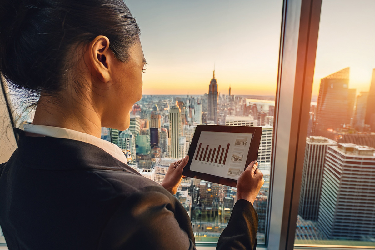 Real estate agent reviewing market reports on a tablet with the New York City skyline in the background, warm afternoon light, professional office setting.